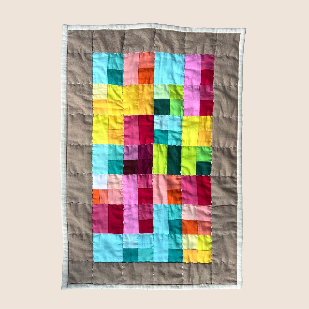 A photo a crinkly and colorful quilt. It’s made up of 28 color coded squares each encoding a book read, listed in 7 rows of 4 quilt squares. Small hand made stitches add texture to the quilt.