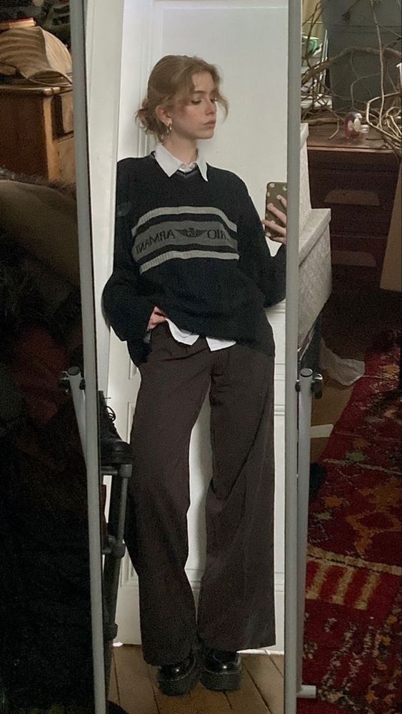 someone wearing a black sweater with grey stripes, a white button down underneath, brown slacks, and black platform creepers. they have their hair done up in a bun, and they look away from the camera.