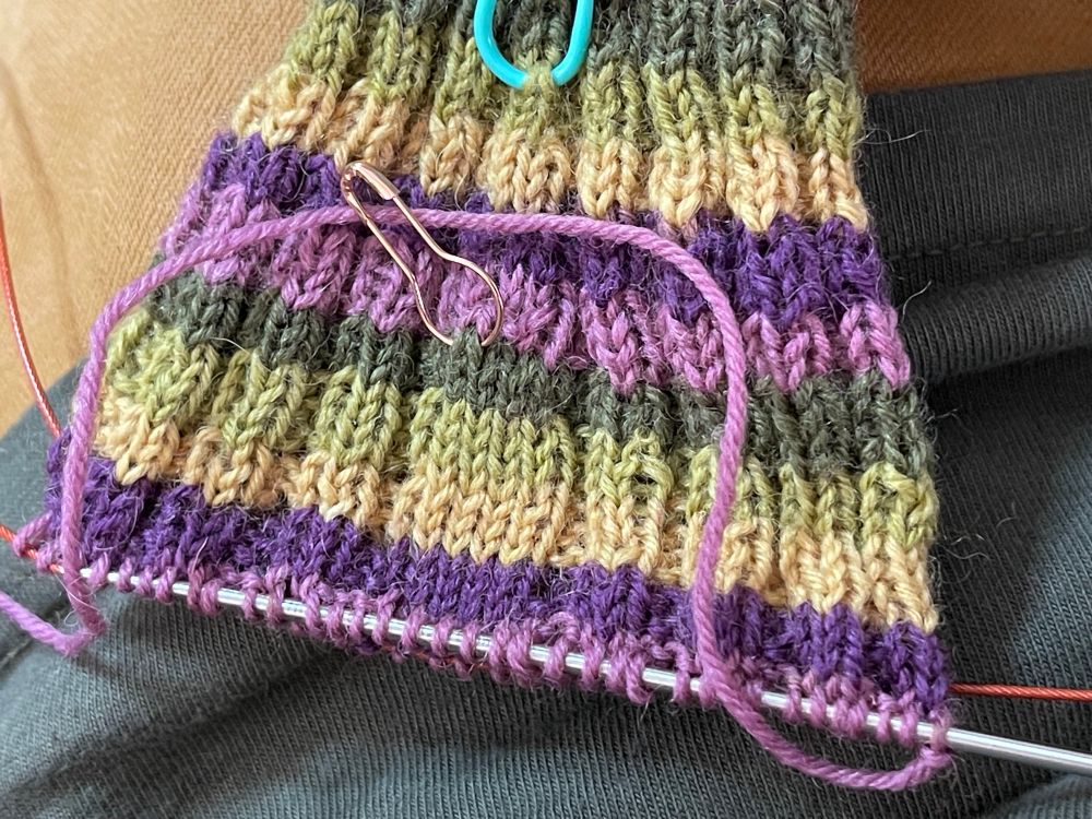 Some ribbed knitting with colorful stripes of lilac, aubergine, lime, yellow and forest green sit on thin knitting needles. It looks like it might be a sock. 