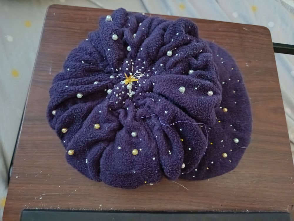 Une casquette gavroche violet foncé avec des perles plus ou moins grosses allant du blanc au doré, du jaune à l'argenté. Vue de dessus et à l'endroit