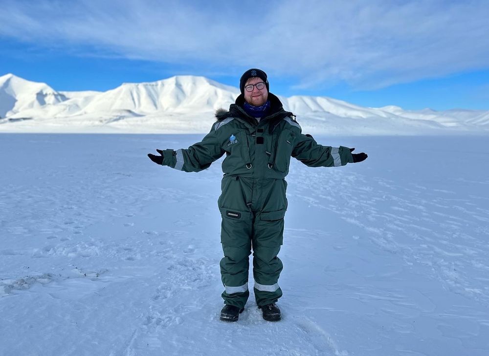 Dylan stood with arms open on snowy ground in Svalbard with snow covered mountains in the background.