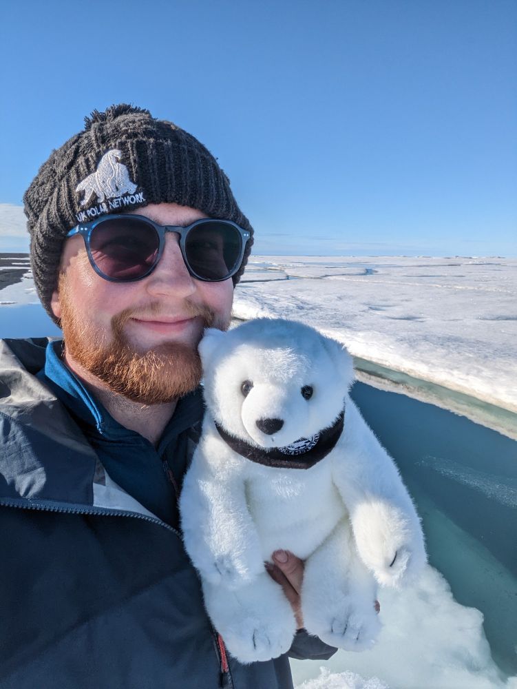 Dylan holding a plushie polar bear while stood on sea ice in Greenland.