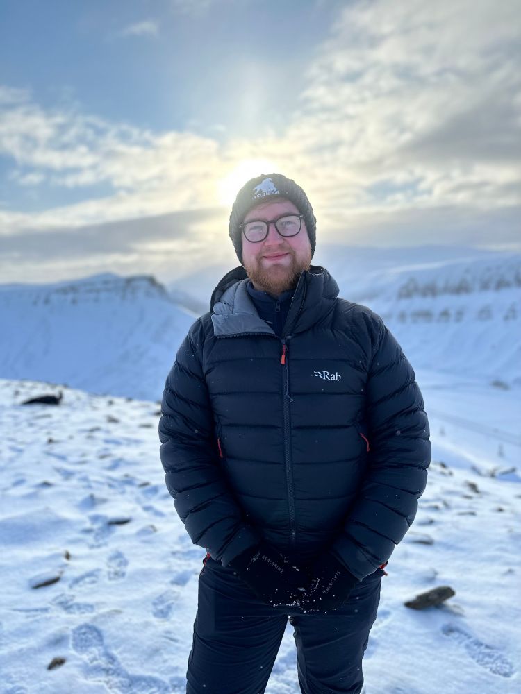 Dylan stood on top of a snow covered mountain in Svalbard with the sun setting behind him.