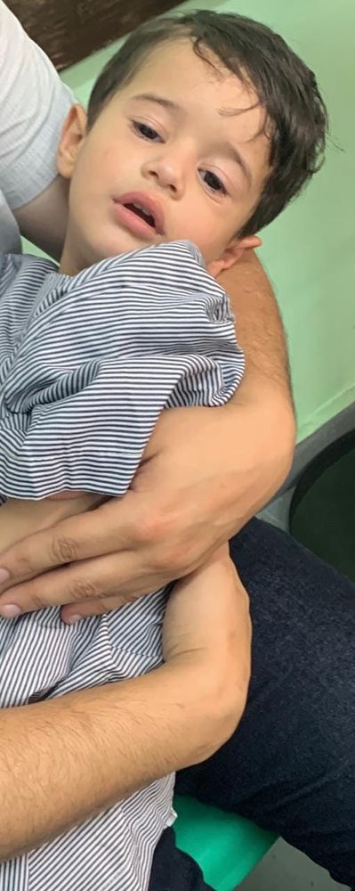 Omar, a 3-year-old Palestinian boy, wearing a hospital gown and being held in the arms of someone out of frame. He looks tired and unhappy.