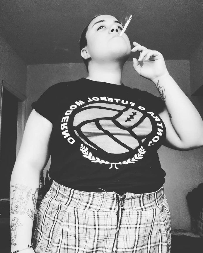 Uma foto minha (não sei me descrever perdão) fumando um cigarro, em pé com uma saia xadrez e uma camiseta escrito "Contra o futebol moderno".