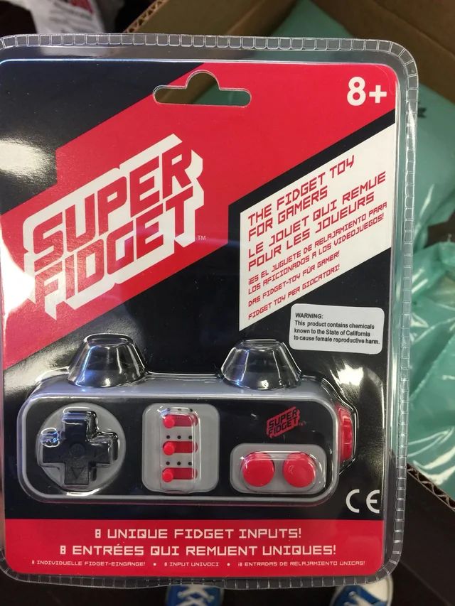 Image of the long-since-discontinued Super Fidget sold at GameStop around 2017, taken from a r/GameStop post. The toy has a d-pad, two plain buttons, and three switches each with three set positions (left, middle, and right) visible on the front side, two joysticks visible on the top (which can be pressed to click like most modern joysticks), and a toggling button visible on the right side. Not shown is a mousewheel on the lower side, a trackpad and trackball (both of which click) on the back, and a place to attach a wristband on the left side.
