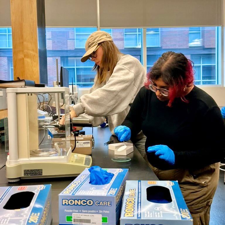 Two students in a lab at Ontario Tech’s north campus. They are standing next to a table working with lab equipment. The student toward the front of the frame is wearing blue latex gloves. 