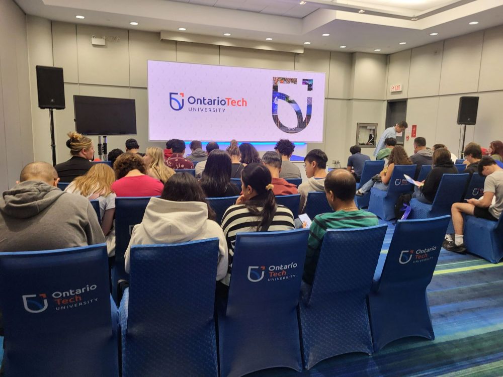 Prospective students fill the seats in a room facing forward. There is a large screen at the front with the ontario tech logo on it. Several chairs have the ontario tech logo on the back. 