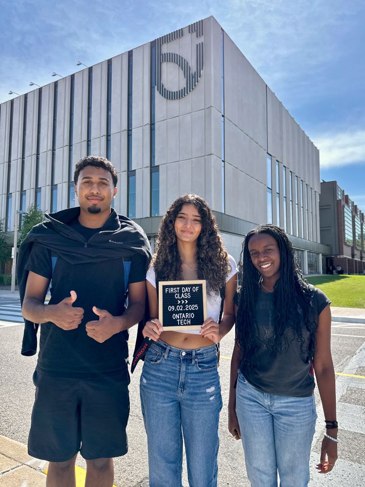 A student at centre holds a small letterboard that reads First Day of Class, 09.02.2025, Ontario Tech. She is standing with two other students in front of Shawenjigewining Hall. The student at left has his thumbs up. 
