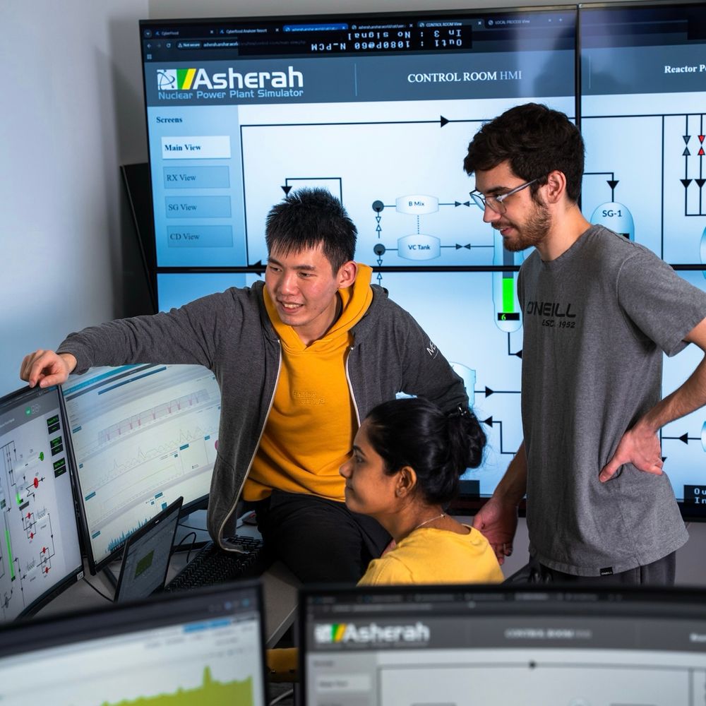 Students in a cybersecurity lab at Ontario Tech University's north campus. Two students are seated in front of computer screens and one student is standing behind them looking toward the screens. There are large TV screens displaying information on the wall behind them.