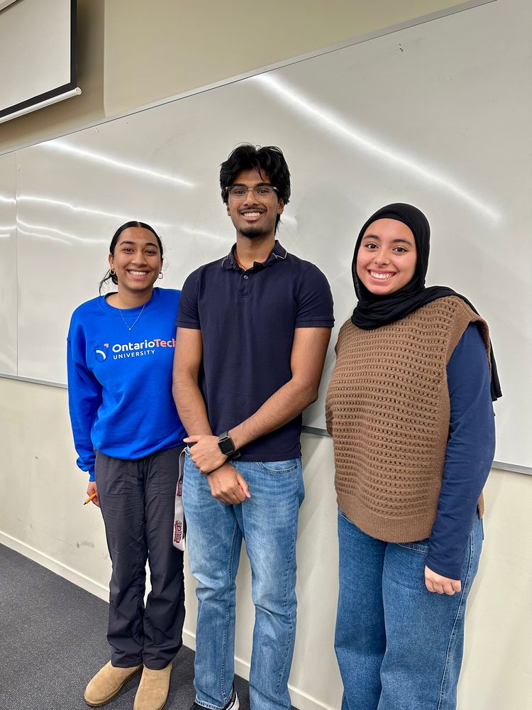 Three students standing at the front of a classroom posing for a photo with a large whiteboard behind them.