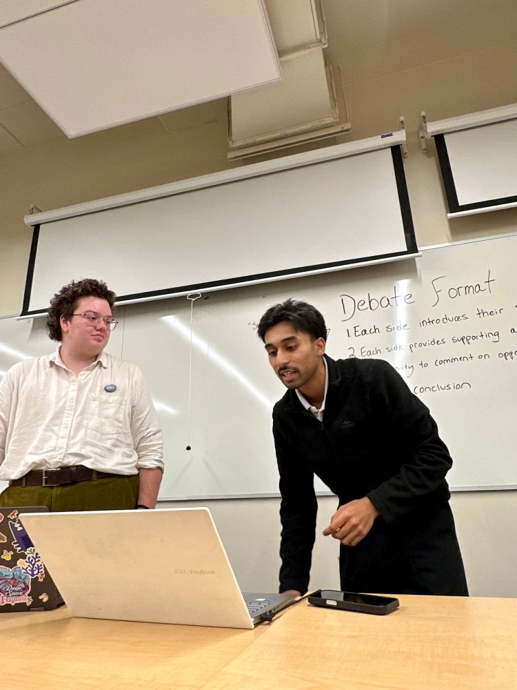 Two students standing at the front of a classroom presenting their points in a debate. The student on the right is looking down at a laptop on a desk in front of him. There is a whiteboard in the background with information on the debate format written on it.