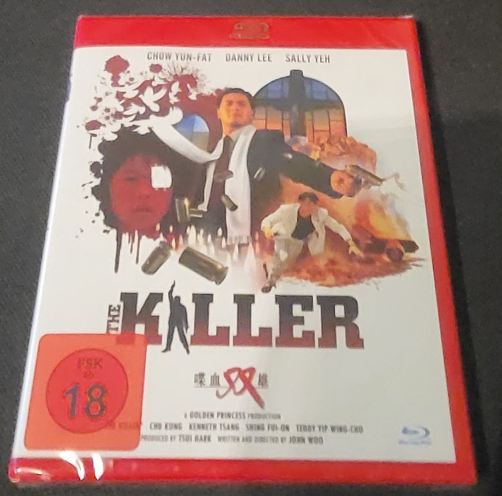 Blu-Ray Cover vom Film The Killer. Zu sehen sind Szenen aus dem Film. In der Mitte ein asiatischer Mann im Anzug mit einer Pistole. Auf der linken Seite eine Frau mit einem Verband über dem Auge. Auf der rechten Seite ein Mann in gestreiften Anzug und eine brennendes Auto.