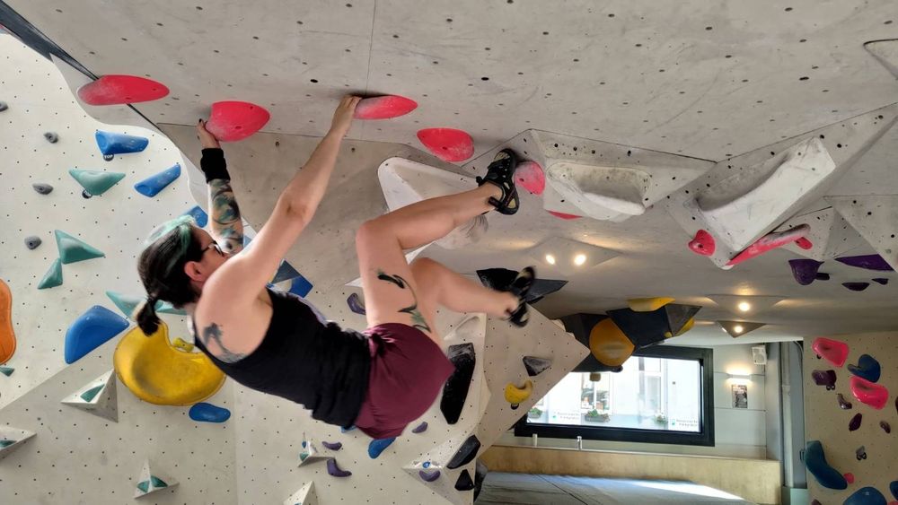 2DKiri at a bouldering gym. She is holding onto ceiling holds with her hands and feet.