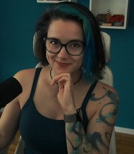 2DKiri sitting at her desk in her studio smiling. She's holding an arm underneath her chin. She's wearing a teal spaghetti strap top.