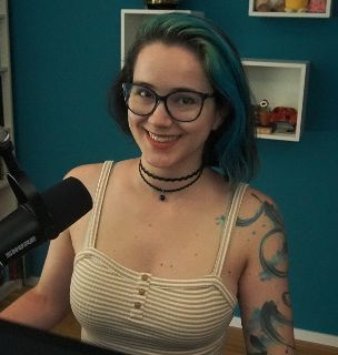 2DKiri sitting at her desk in her studio smiling. She's wearing a white and brown striped spaghetti strap top and a double choker.
