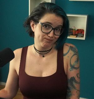 2DKiri sitting at her desk in her studio shrugging and looking thoughtful. She's wearing a maroon tank top and a black choker.