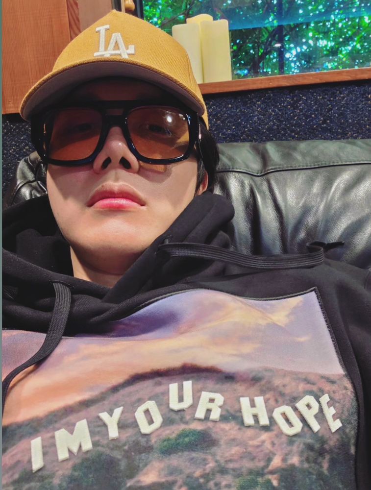 Hoseok wearing glasses, a yellow LA ball cap, and a hoodie that says I’m Your Hope