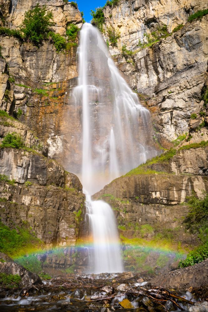 A towering white waterfall cascading over a steep rocky cliff with multiple levels. A vibrant rainbow shines at its base. 