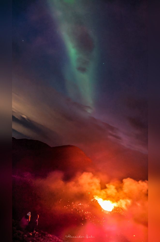 Two people sit on a steep hill directly before an erupting volcano. The landscape glows bright orange, with gases filling the scene. Overhead, a long and green streak of the northern lights cuts across the mostly clear blue night sky. 