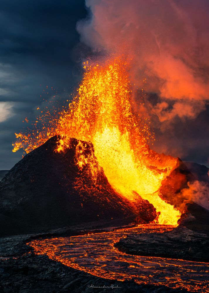 An erupting volcano. A black spatter cone with a gap in the front has lava fountaining out, hundreds of feet into the air, smacking against the cone, and flowing along a snaking lava river toward the camera. A plume of gas rises into the air with mostly cloudy sky in the background.