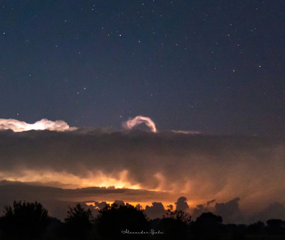 A photo of a distant thunderstorm displaying an above anvil cirrus plume in the center of the frame - a narrow loop/curl in the clouds. The thunderstorm clouds in the bottom half of the image are illuminated yellow-orange by in-cloud lightning. Above the storm is a starry night sky.