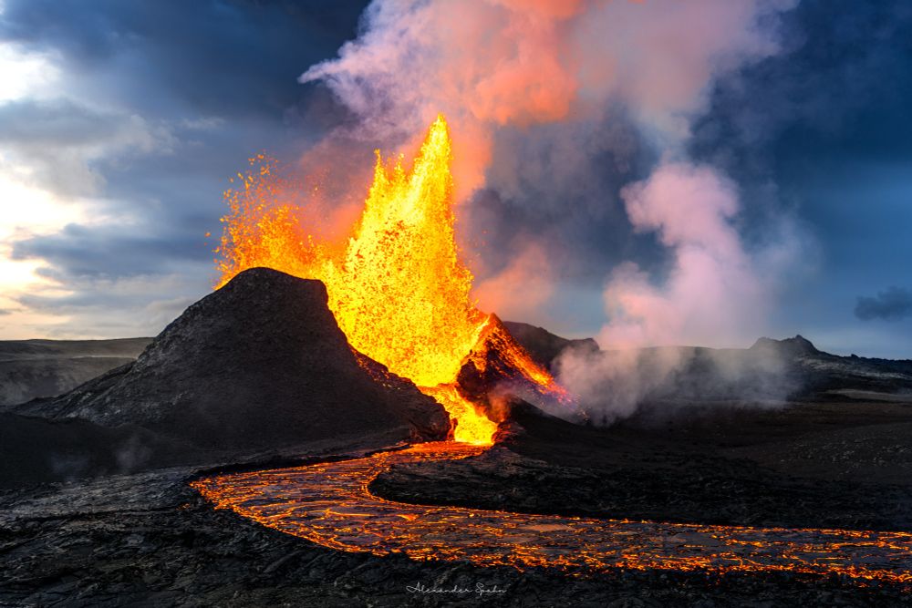 A photo of a volcano erupting. Vibrant yellow-orange lave erupts from a black spatter cone, with white clouds of gas rising into the mostly cloudy sky and a river of lava snaking toward the camera. 