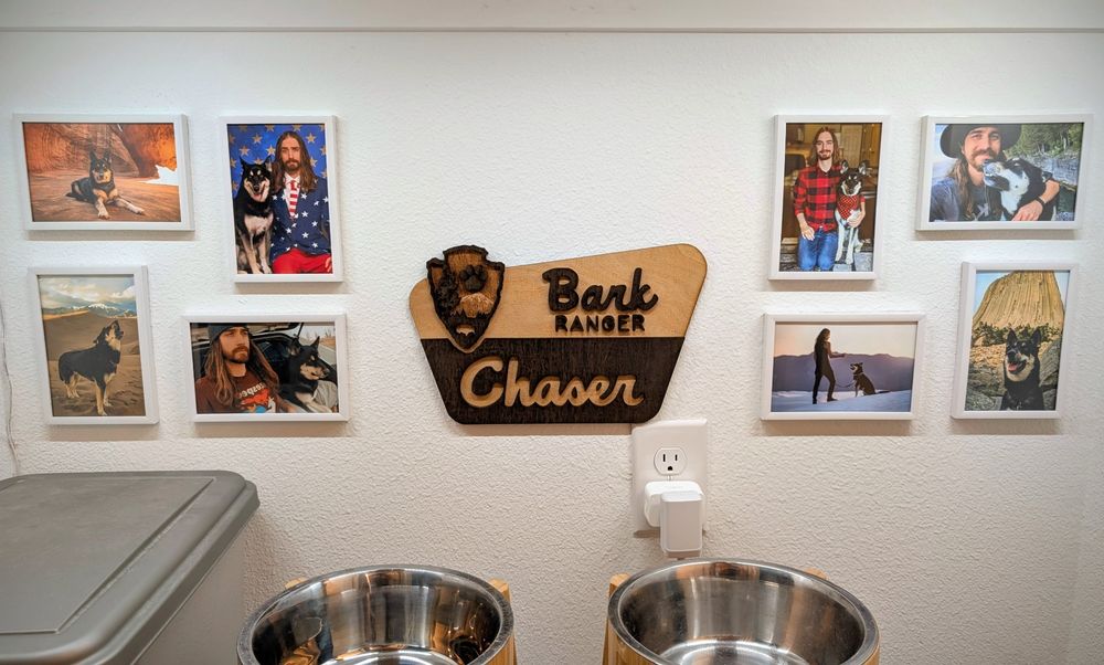 A picture of my dog's feeding area, with bowls on stands. LED lights illuminate the space, and there are 8 small, framed photos of Chaser/Chaser and I on the wall behind the bowls, with a sign in the middle in the shape of a National Park sign that says "bark ranger." 