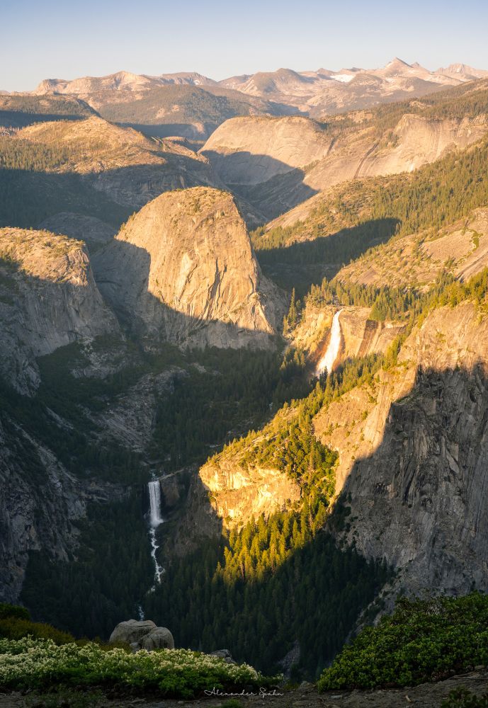 A portrait photography of Vernal Falls, Nevada Falls, and Liberty Cap in Yosemite with the Sun having nearly set on the horizon off the the left (out of frame). Distant mountains with a little bit of remaining snow point into the clear blue sky overhead at the top of the frame. A patch of greens and white flowers dot the foreground at the bottom of the frame.