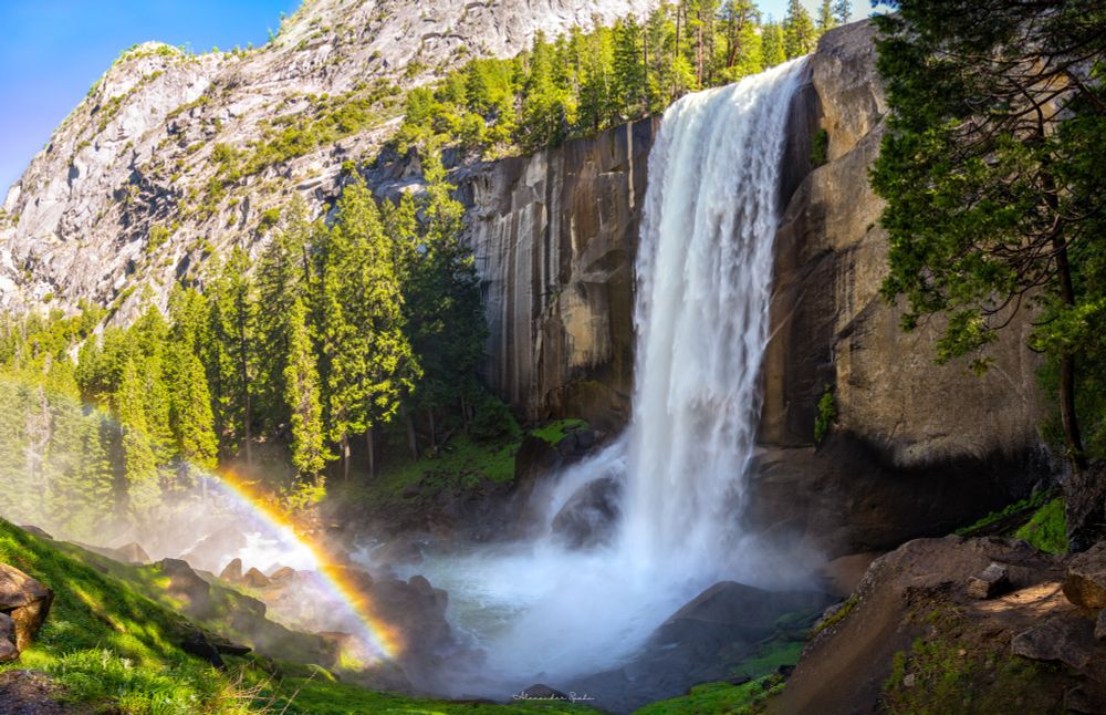 A photo of Yosemite's Vernal Falls. Water plunges over a tall rocky cliff on the right, creating a mist at the surface. Sunlight hitting the mist forms a rainbow on the left side of the image. The rocky landscape is partially covered in tall green trees. Small patches of blue sky are visible over the rocky landscape.