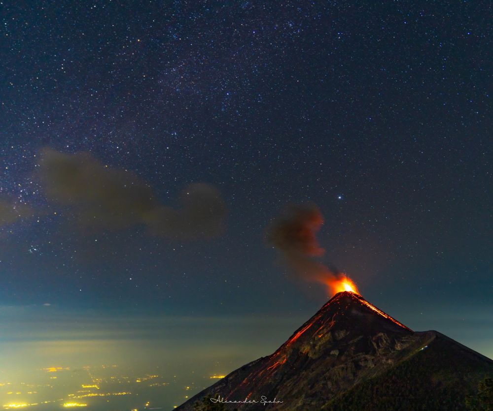 A volcano erupts thousands of feet above the surrounding landscape, with golden city lights illuminating a layer of haze., and a clear starry night sky above.
