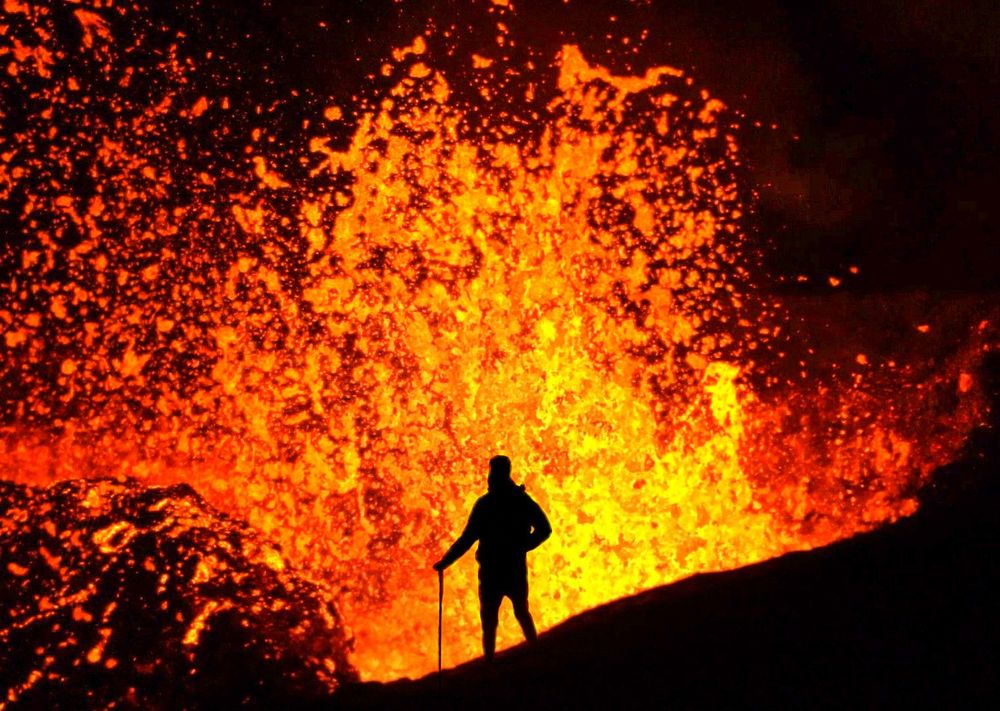 A photo of me standing on a hill immediately before an erupting volcano. Via zoom / lens compression, it appears as if my silhouette is right on the edge of the volcano, but I am a safe distance away from the bright, vibrant, orange-yellow lava propelled above the Earth's surface. 