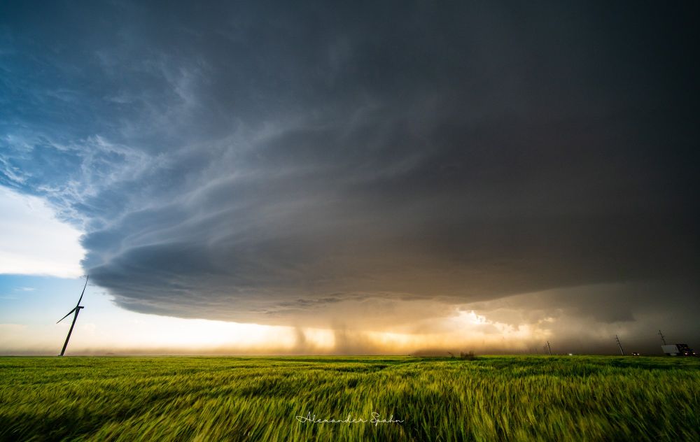 A supercell thunderstorm with a pancake stack appearance hovers over fields of tall, green grass. A lone wind turbine stands tall on the left, where there are clear blue skies. A highway is on the right, where the storm's core is dumping rain and large hail in a darkened scene. Brown dust/smoke is being pulled off the ground and up into the center of the storm due to its strong inflow. Behind the dust/smoke is the almost-setting Sun's golden light. 