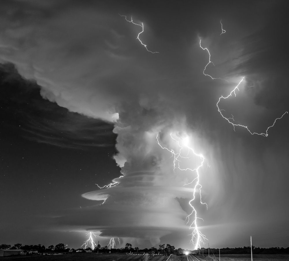 A black and white photo of a low precipitation (LP) supercell at night. The supercell's mesocyclone is remarkably sculpted into a 'barber pole' like appearance, with three lightning bolts striking the ground around it, and several intracloud bolts as well. To the left of the storm's towering updraft is a clear, starry night sky. 