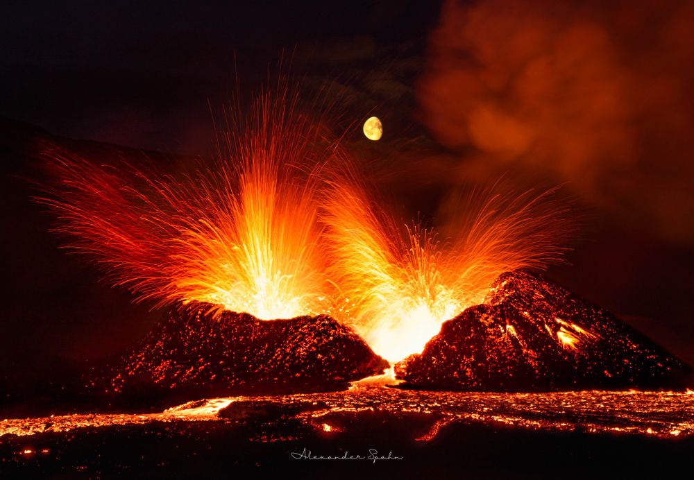 A gibbous Moon sits directly above an erupting volcano, with lava fountaining in all directions and a lava pool sitting out front.
