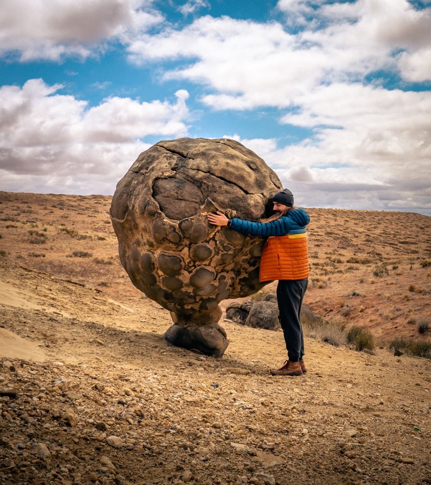 A photo of me hugging a large, natural, spherical rock on a rocky pedestal out in the middle of nowhere of a desert landscape in Arizona.