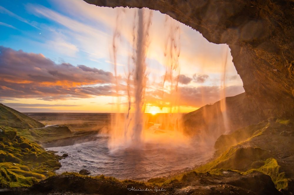 A photo looking toward the Sun as it rests on the horizon at sunset. The photo was taken from behind a waterfall, which is centered in the frame, looking out at the dynamic and vibrant sky through the water. The sky has a golden glow around the Sun toward the right of the image, while blue skies persist toward the left. A tiny crescent Moon is visible in the blue of the sky. In the foreground are rocks/slopes covered in green moss, with the rocky cliff from which the water falls wrapping around the right side of the frame. Seljalandsfoss, Iceland (2021)