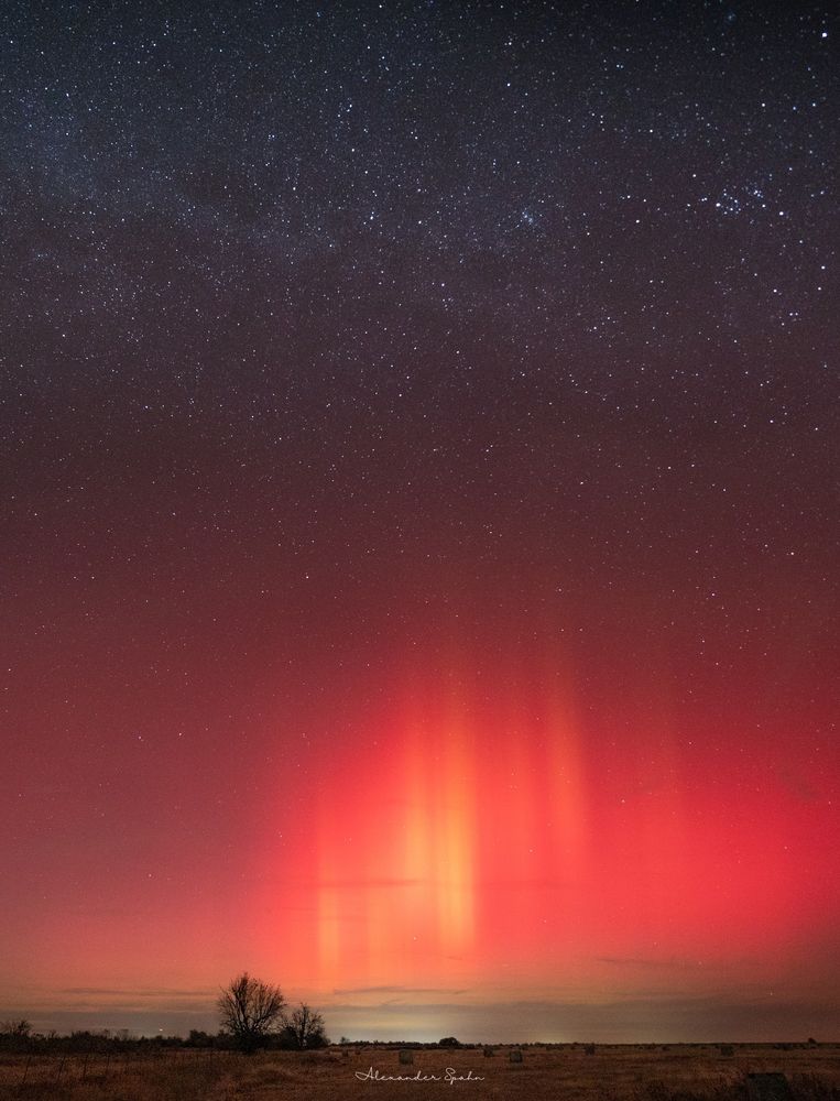 A photograph of tall, vibrantly red-orange aurora pillars on the horizon. The foreground is a brown farm field with a few bare trees and hay bales. Above the aurora is the faint band of the Milky Way. 