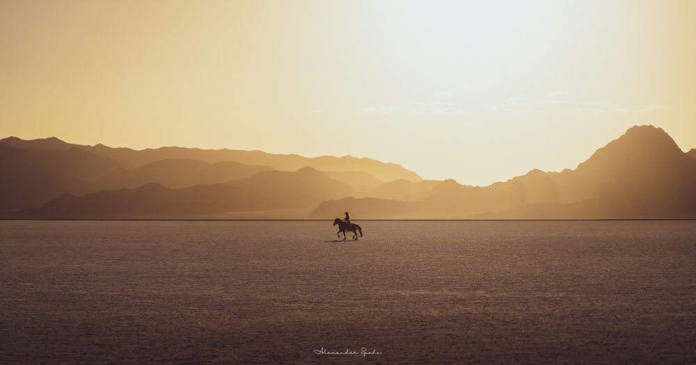 A woman on horseback rides across the Bonneville Salt Flats at sunset. The scene is hazy and all is golden, with the Sun's rays piercing through the haze along the more distant mountain peaks.