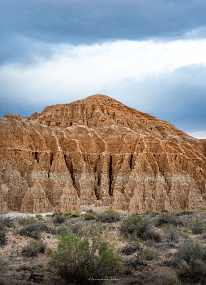 A cathedral of eroded bentonite clay before a desert landscape dotted with dry shrubs and below a clouded, stormy sky.