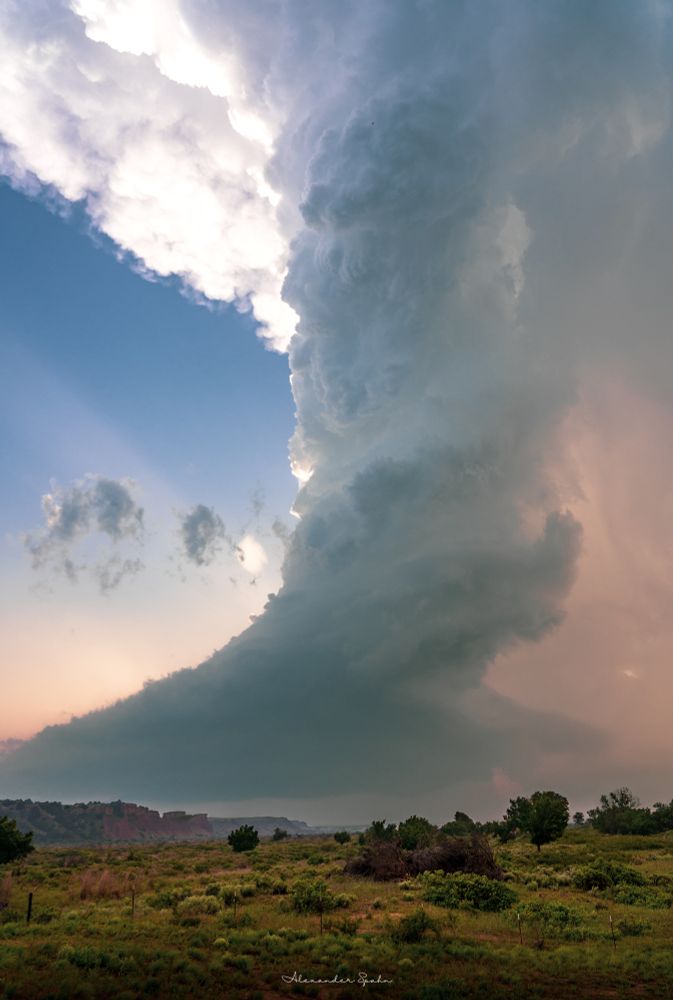 A ridiculously tall LP supercell towering over a northwest Oklahoma landscape with red cliffs in the distance. The Sun appears to be setting behind the storm. 