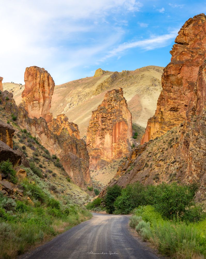 A picture looking straight down a gravel road in a canyon bracketed by steep hills with some green vegetation and massive, towering orange pillars of rock.