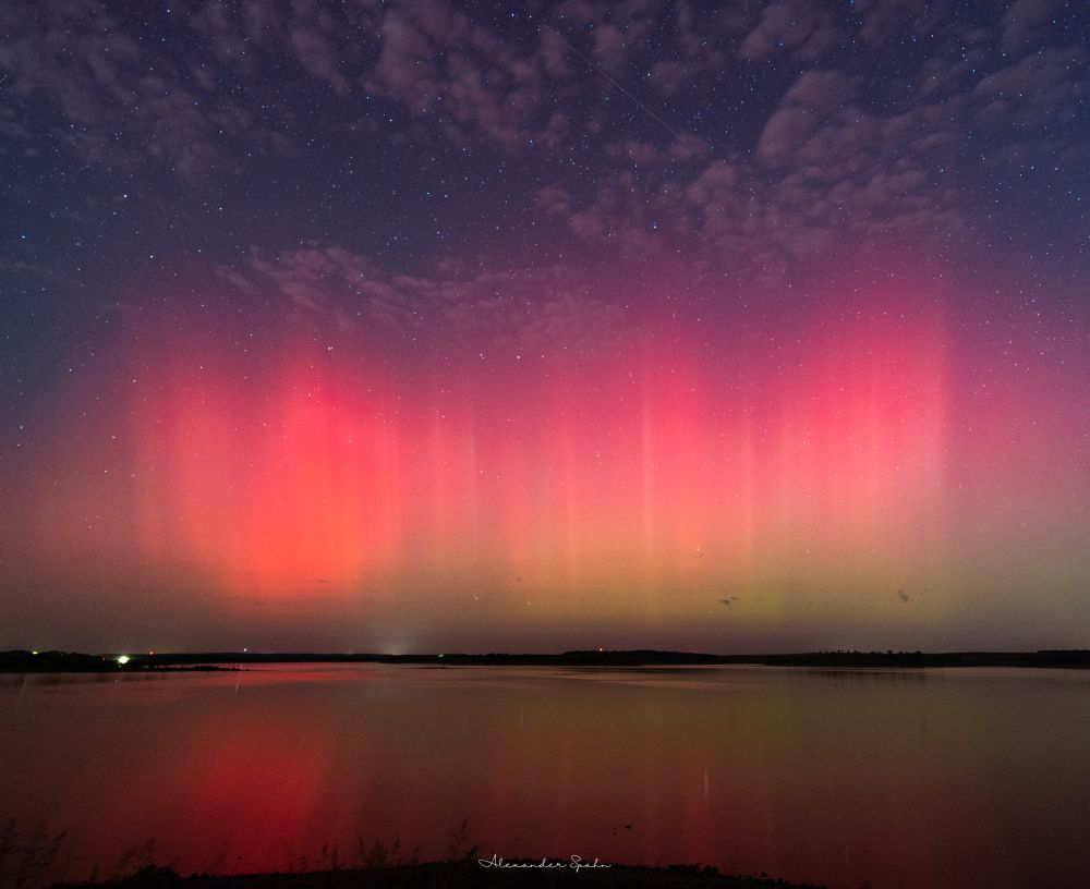 Numerous pinkish aurora pillars with a bluish-purple sky and spotty clouds over relatively still waters of a large lake. 