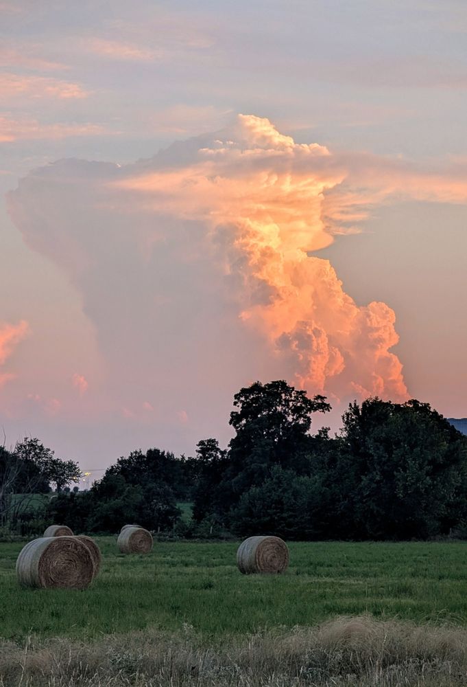 A sunlit, towering storm in the distance over a field with hay bales.
