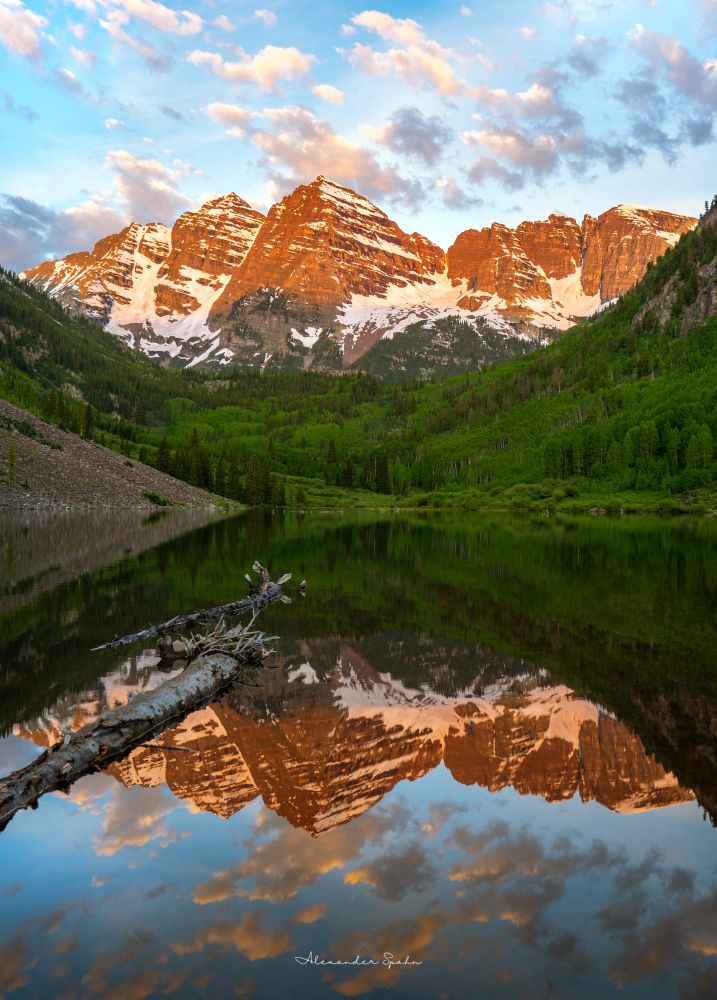 An image taken of Colorado's Maroon Bells from the still lake before it. In the foreground is the glass lake mirroring the landscape and sky above. A long log on the water reaches out from the left of the frame toward its center. On the other end of the lake is a lush green forest, with the forest stretching up the steep hills that bracket the image. Sitting right in the center of those hills are the Marron Bells - mountain peaks - partially coated in snow, and bathed in the saturated orange glow of the just-rising Sun. Overhead are blue skies with numerous puffy white clouds, some of which are still in the shadow of other mountains. 