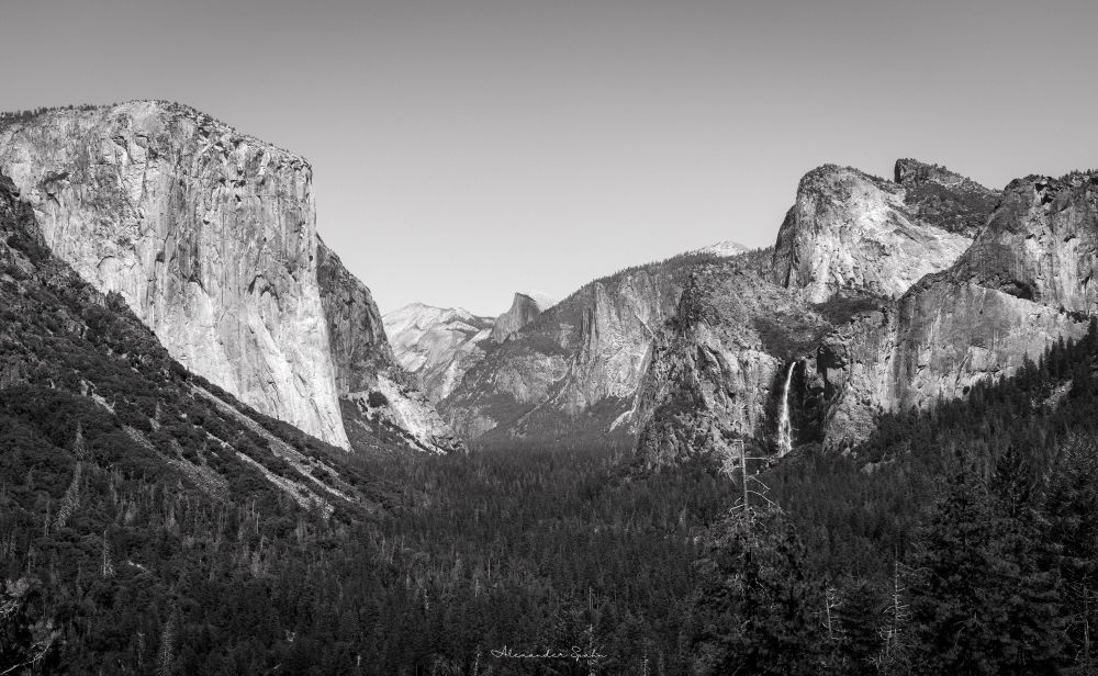 A black & white photo of Yosemite Valley from the Tunnel View. Large granite domes bracket the frame, with Bridal Veil Falls crashing down into the valley on the right and numerous trees below.