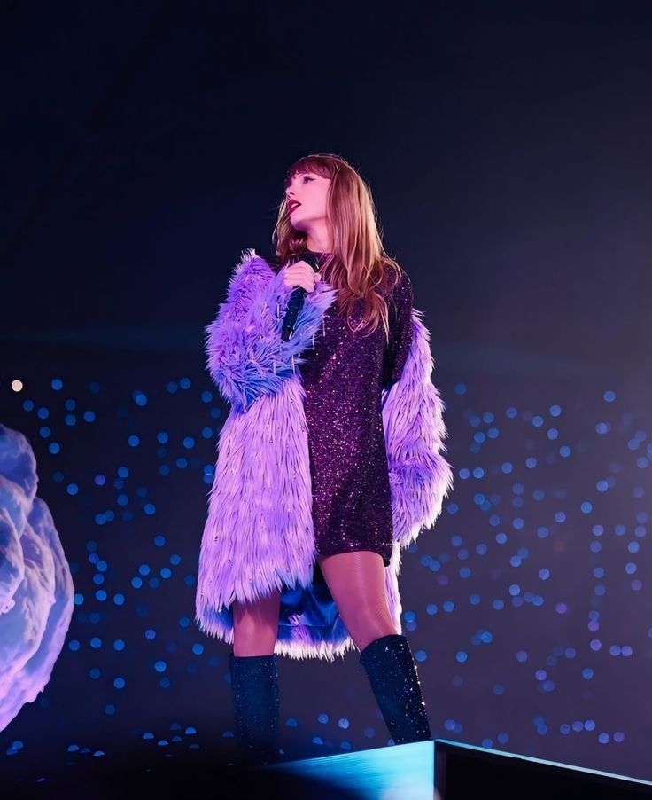 Taylor Swift looking over her shoulder while performing at Eras Tour wearing dark purple Midnights shirt and purple Lavender Haze jacket.