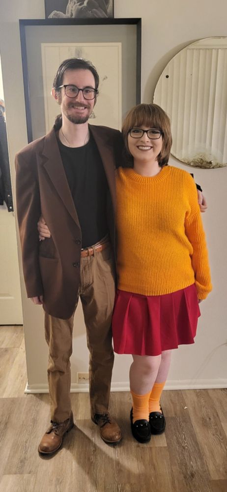 The poster, Steph (right), and their husband, Nathan (left), posing for a picture in their Halloween costumes. Nathan is wearing glasses, a black shirt, a brown blazer, tan dress pants, and brown shoes. He has dark hair pulled back in a ponytail, and he has a goatee. Steph is wearing glasses, an orange sweater, a red skirt, orange socks, and black loafers. They have light brown hair in a bob.