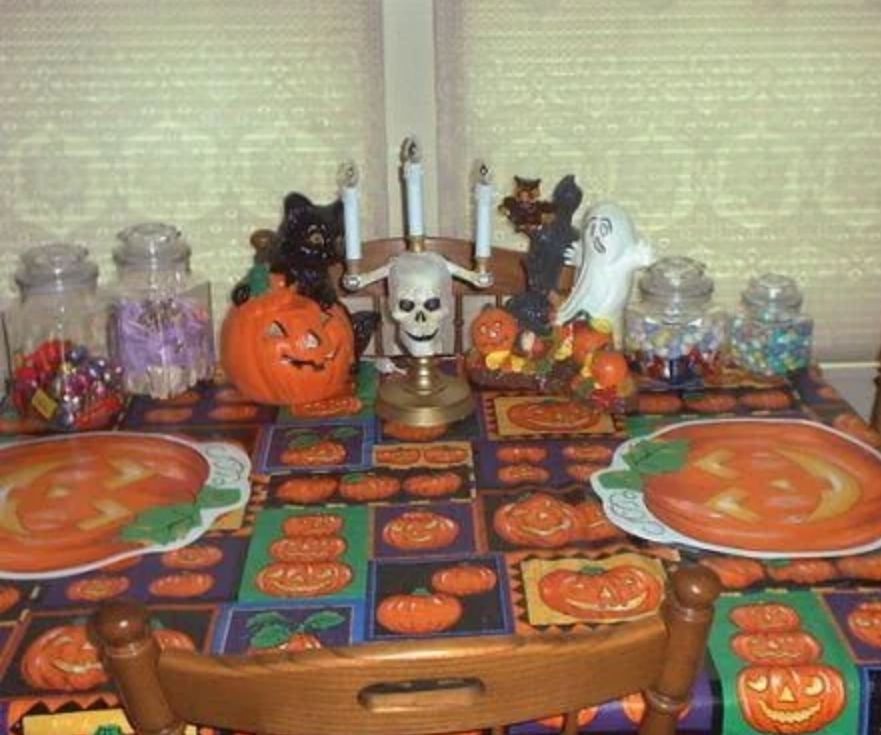 A wooden dining room table and chairs decorated for Halloween. The tablecloth has patches of purple, yellow, green, and black squares with images of jack o lanterns and pumpkins in them. There are four glass jars of candy on the table. In the center of the table is a candlestick with three candles and a skull. On either side are statues, one of a black cat and a jack o lantern, and the other of a ghost, owl, tree, and jack o lantern.
