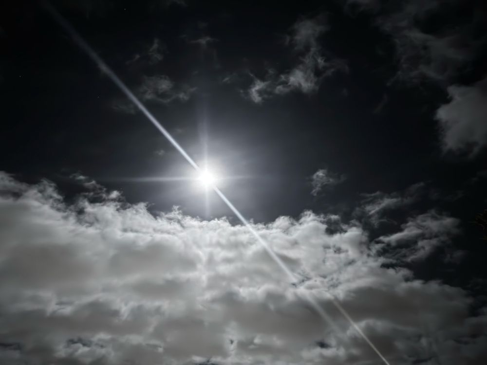 In a black and white photograph, fluffy storm clouds are illuminated by a sparking moon set in the night sky of San Jose, CA.